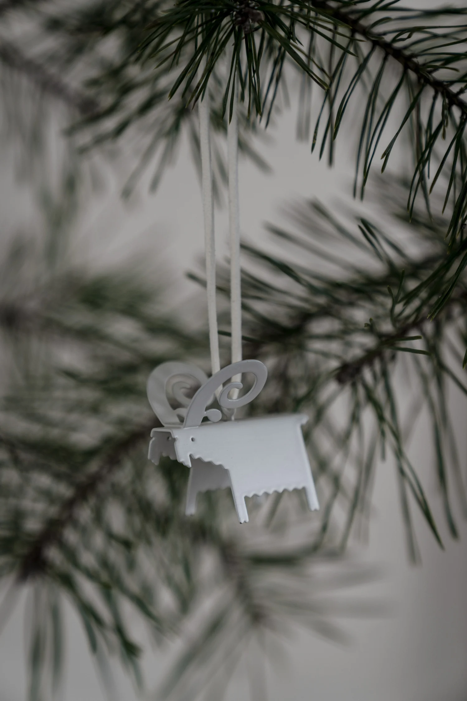 A white metal sheep ornament with curled horns hangs from a pine branch.