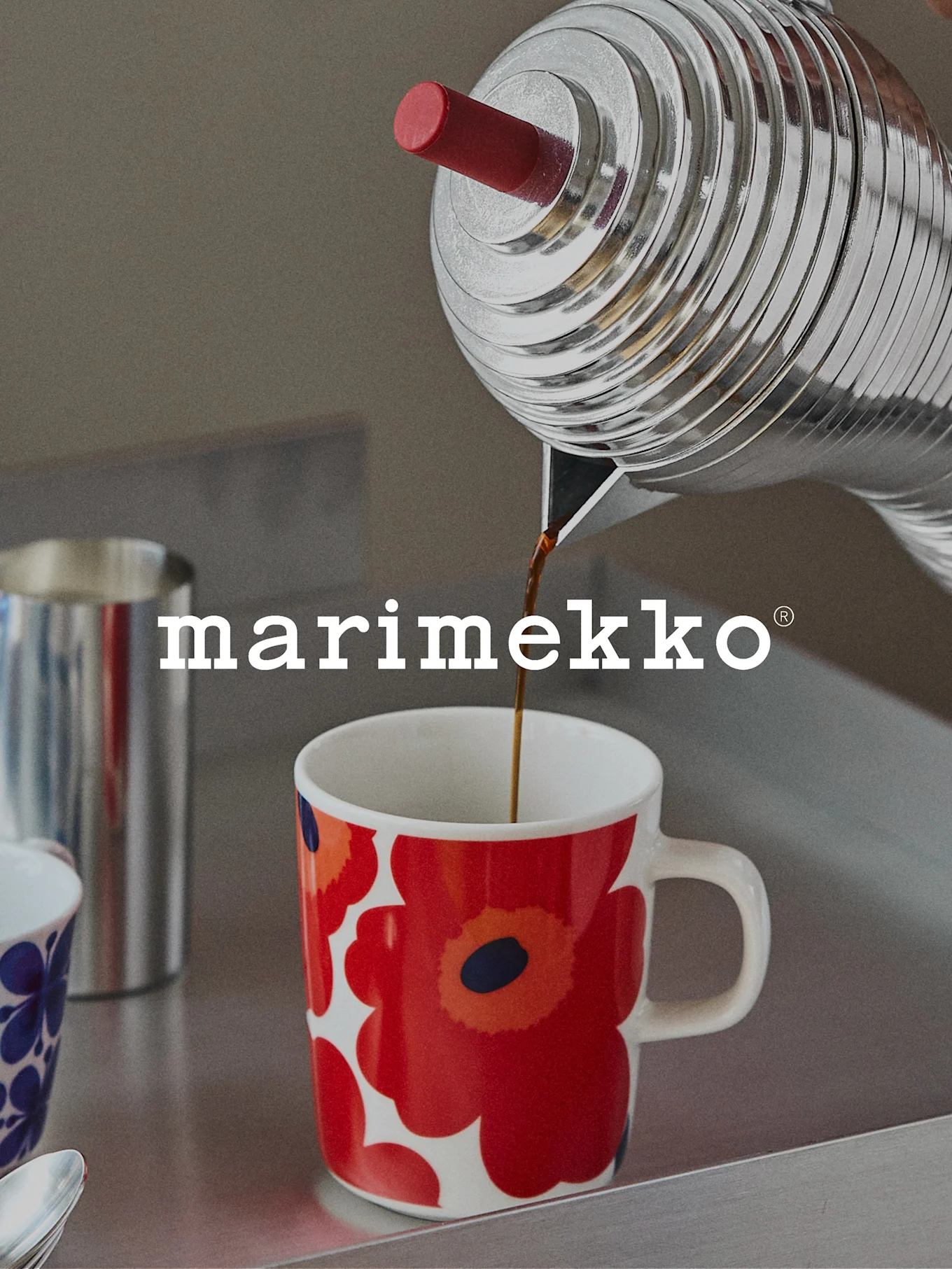 Coffee being poured from a silver carafe into a white mug with a red and blue floral pattern. Marimekko logo visible.