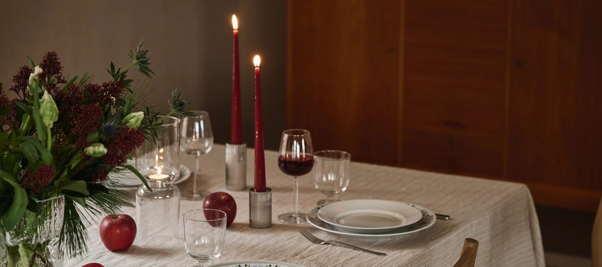 A festive dining table with a floral centerpiece, lit red candles, wine, plates, and red apples on a linen cloth.