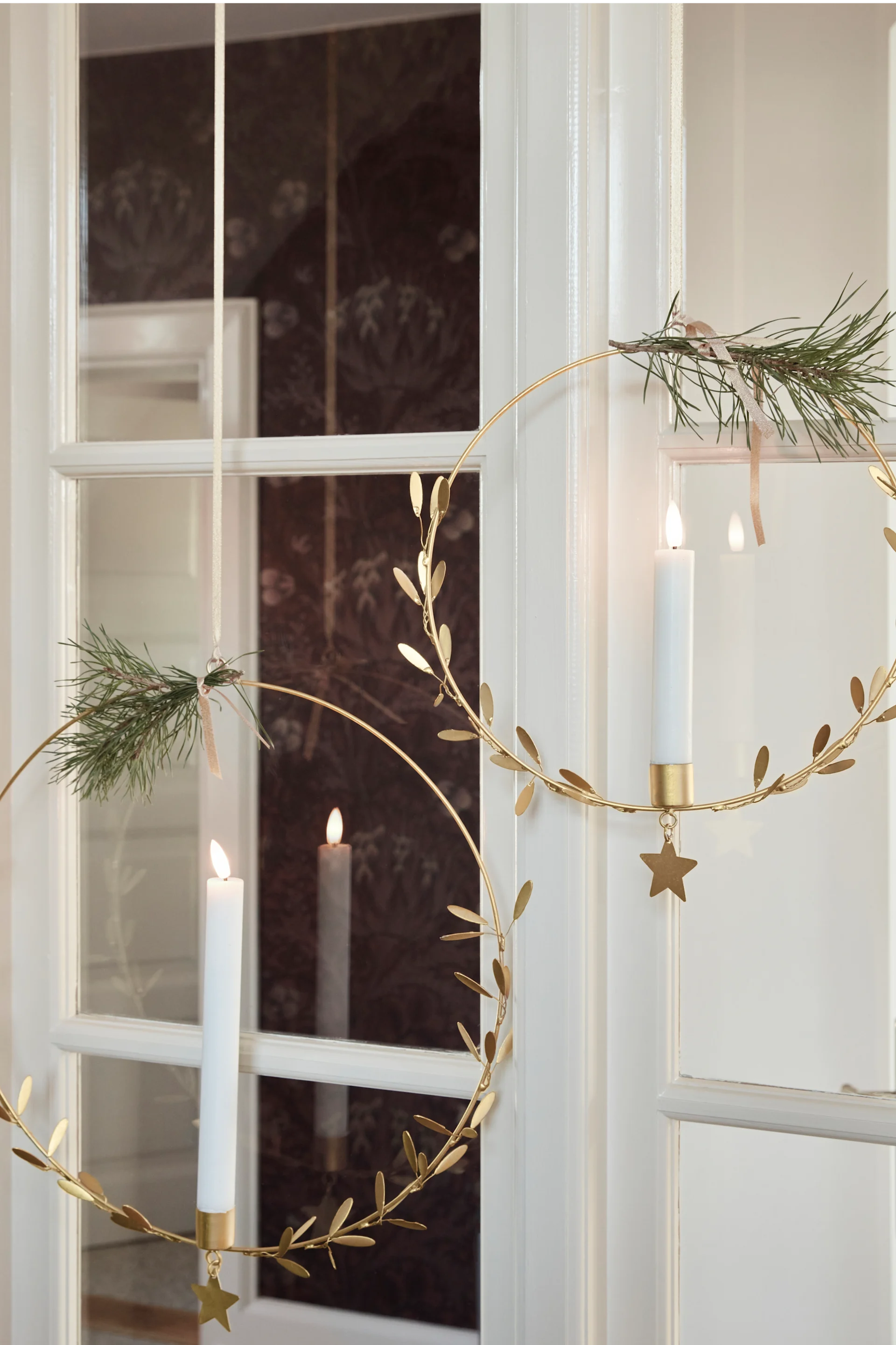 Two golden metal rings, one with a lit white candle and pine sprig, hang against a white door.