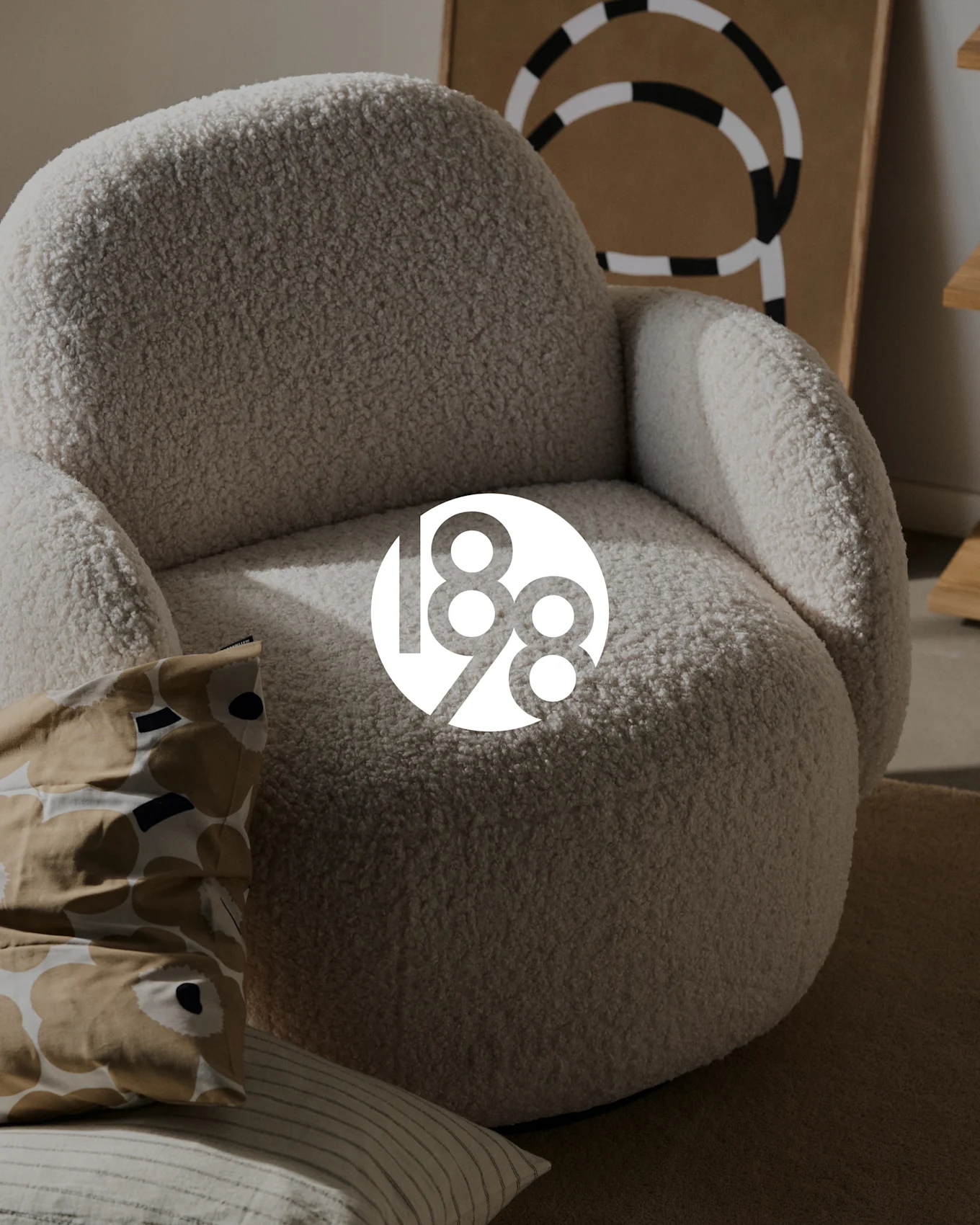 A cozy, cream-colored boucle armchair with a patterned cushion and a striped cushion on a brown rug, with abstract artwork in the background.