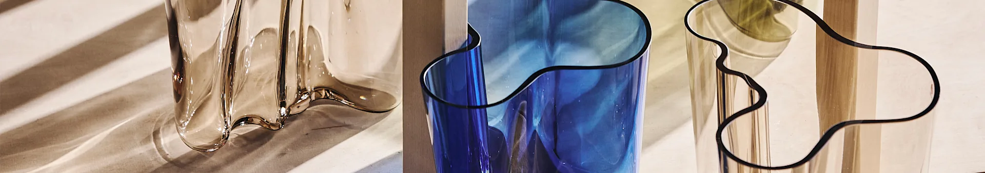Close-up of three wavy, translucent vases: amber, blue, and clear, reflecting striped shadows.
