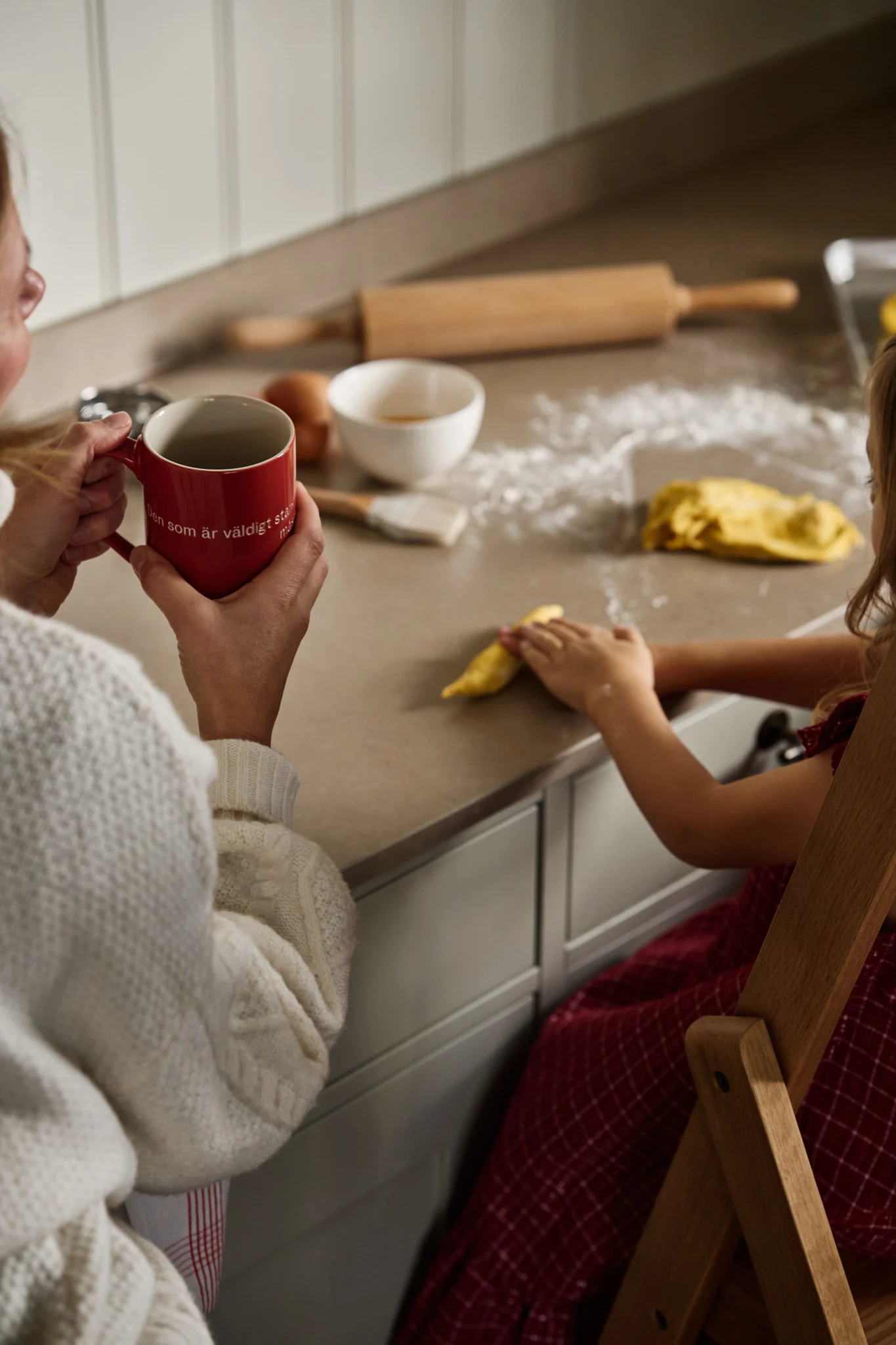 Woman in a cream sweater holds a red mug while a child plays with yellow dough on a floured kitchen counter with baking ingredients.