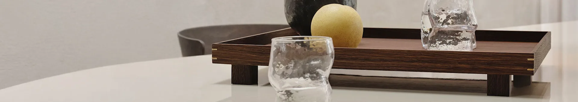 A wooden tray with brass accents holds a light-colored pear and a textured glass. Another glass sits in front on a white table.