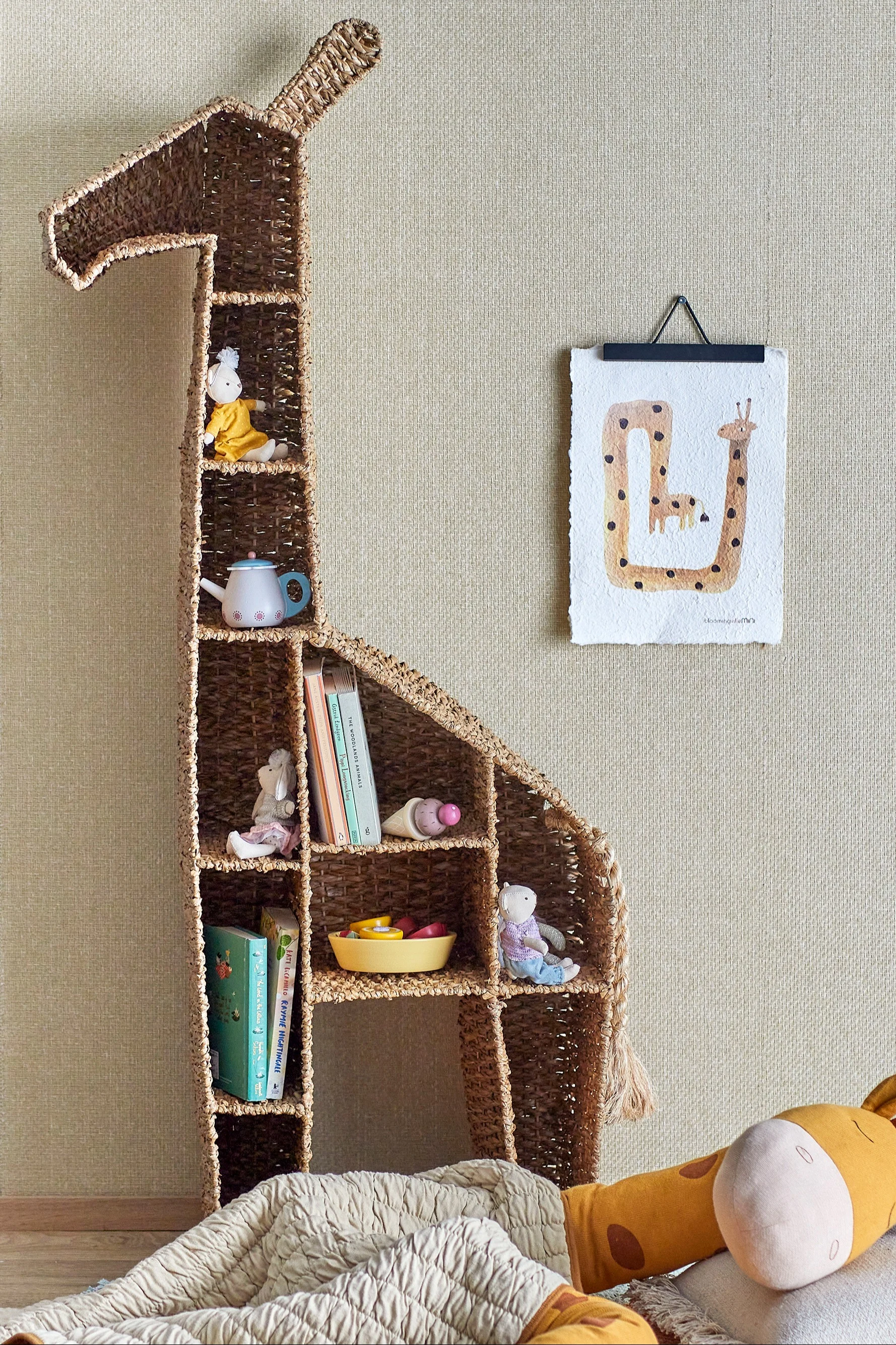 A tall, woven giraffe-shaped shelf unit filled with children's books and toys, next to a giraffe art print, in a child's room.