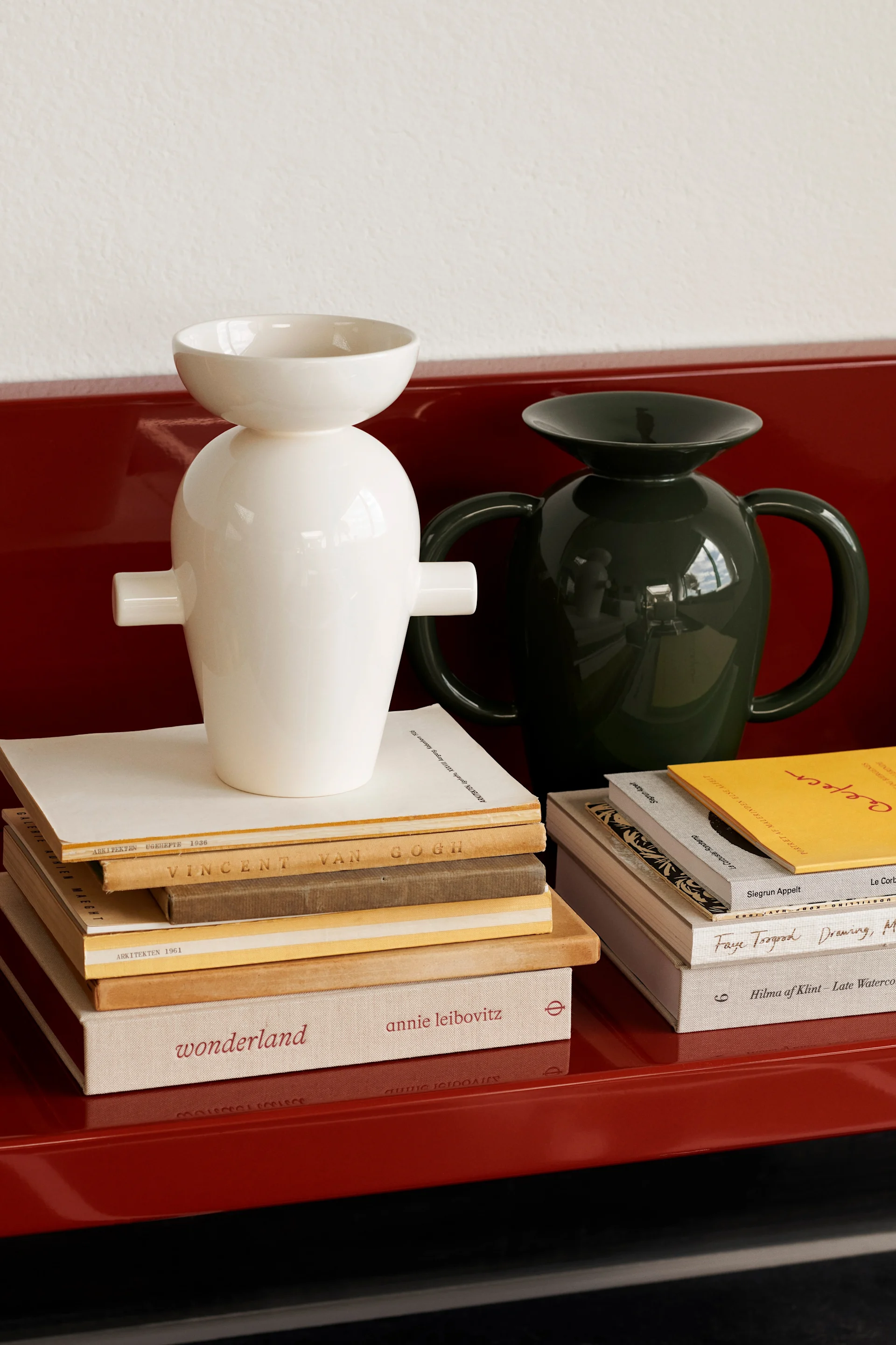 Two glossy ceramic vases, one white and one dark green, on stacks of books placed on a red surface.