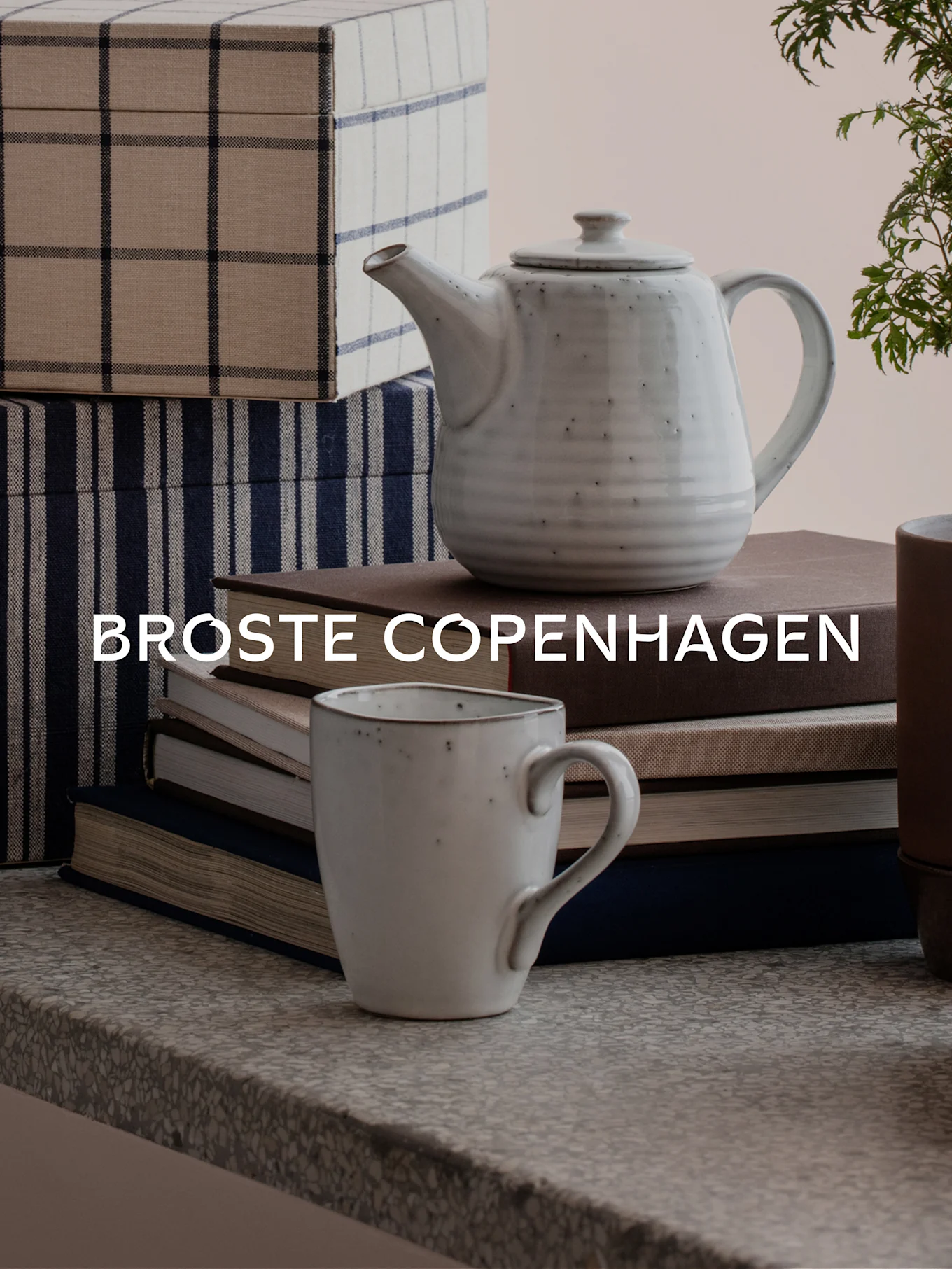 A light gray speckled teapot and mug on books, next to patterned storage boxes. Text: BROSTE COPENHAGEN.