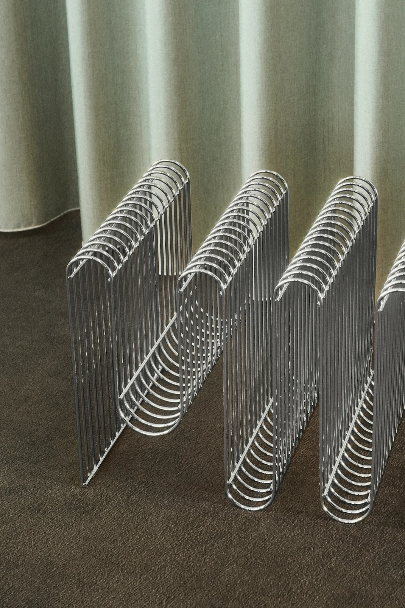 Wavy chrome metal organizer on a brown surface with a green pleated curtain.