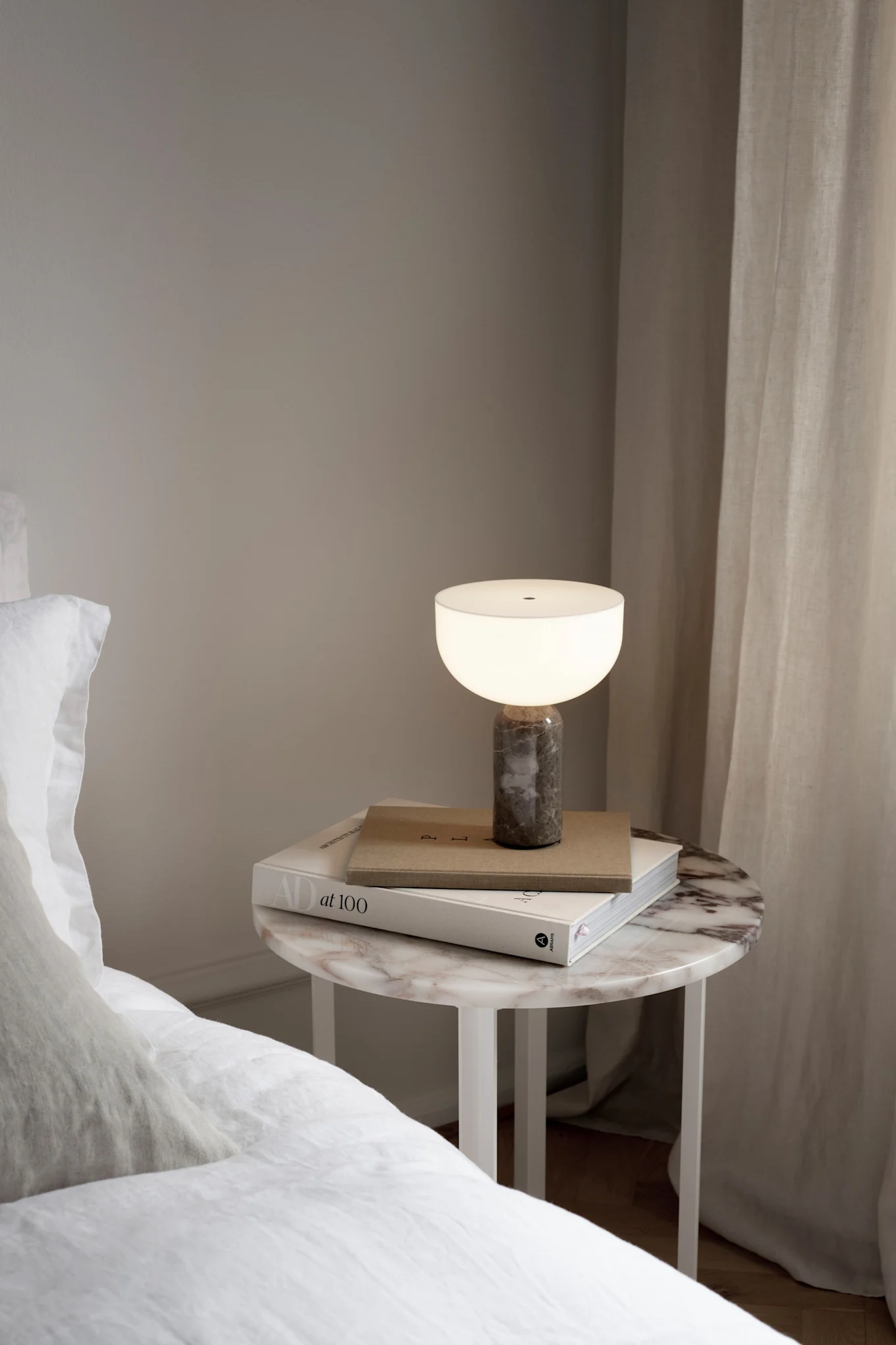 A glowing table lamp with a dark marble base and a white shade on a round marble bedside table with books.