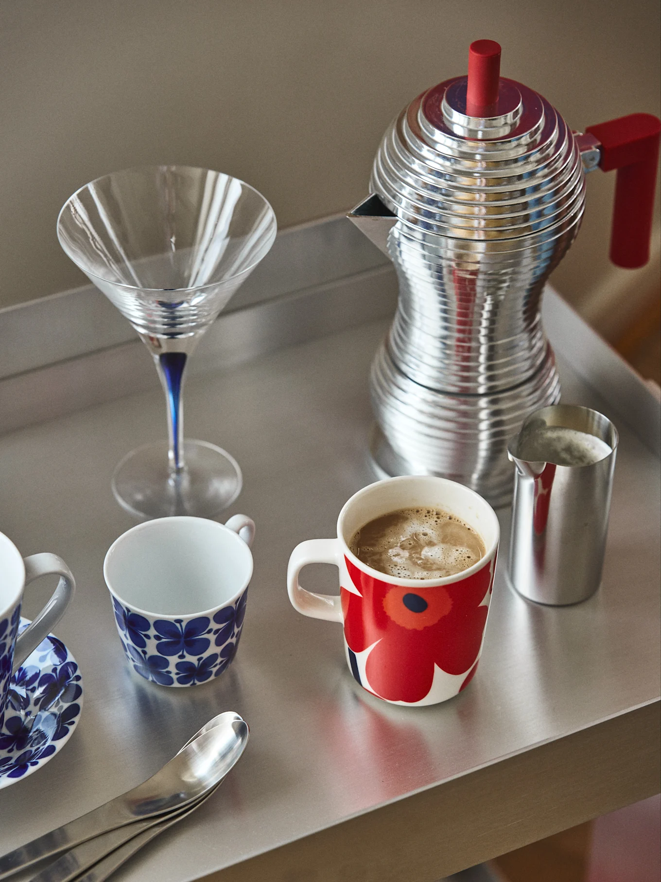 A metal tray holds a red and silver coffee maker, a martini glass, a red floral mug with coffee, a silver milk pitcher, and blue patterned cups.