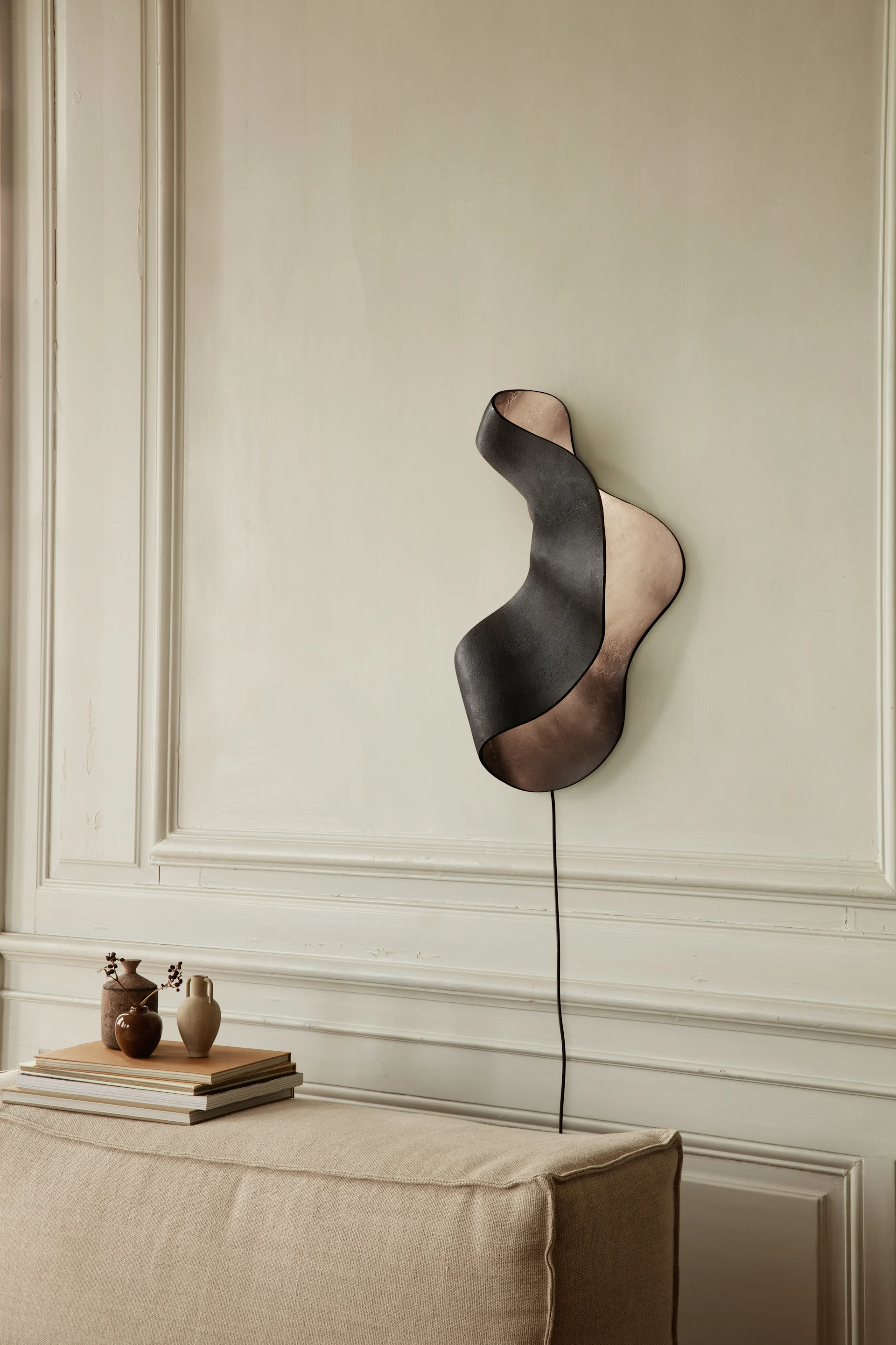 Wavy wall lamp with dark outer and copper inner surfaces mounted on a paneled wall above a fabric bench.