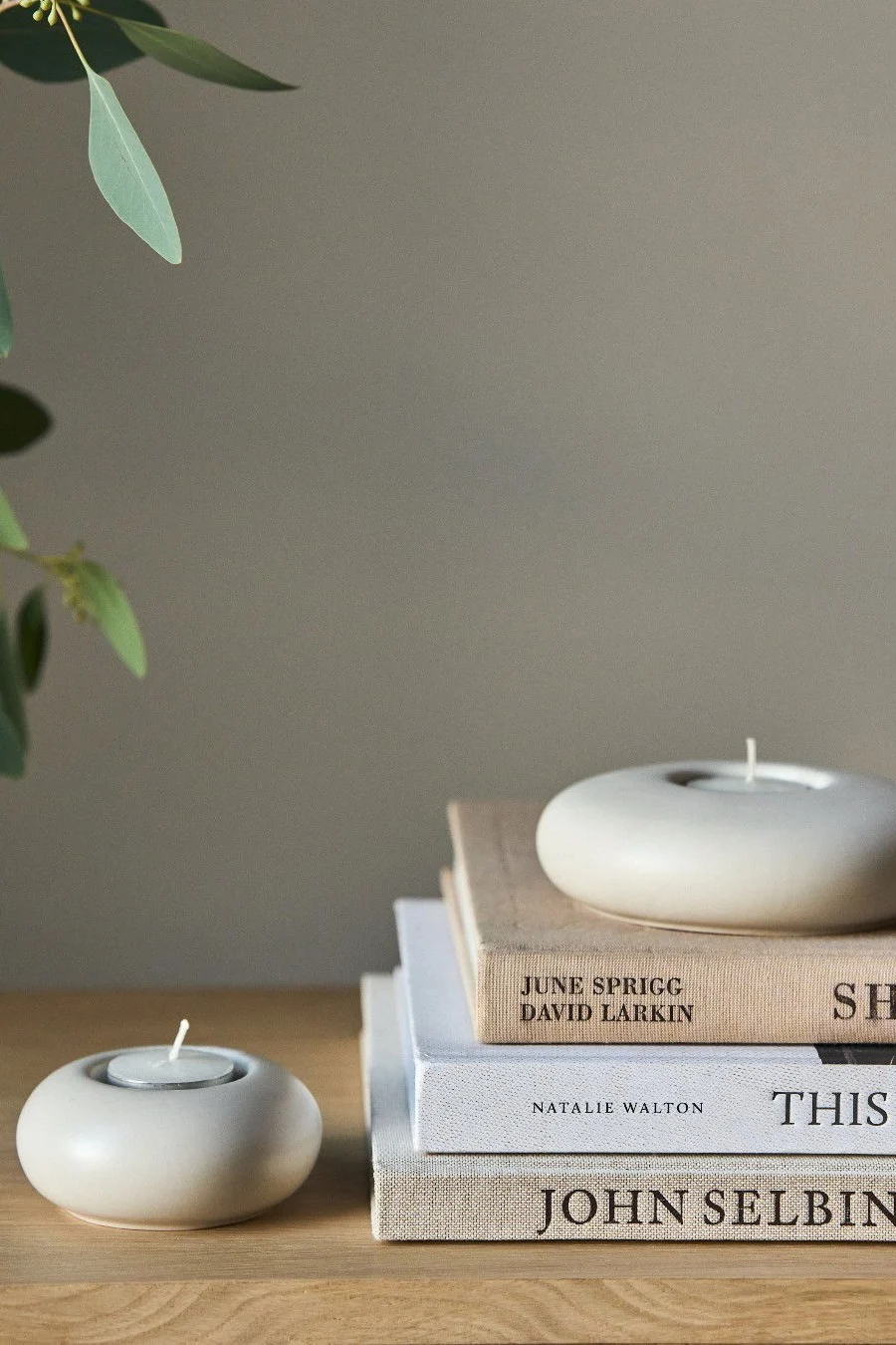 Two round, light-colored ceramic candle holders, one on a stack of books, and a eucalyptus branch.