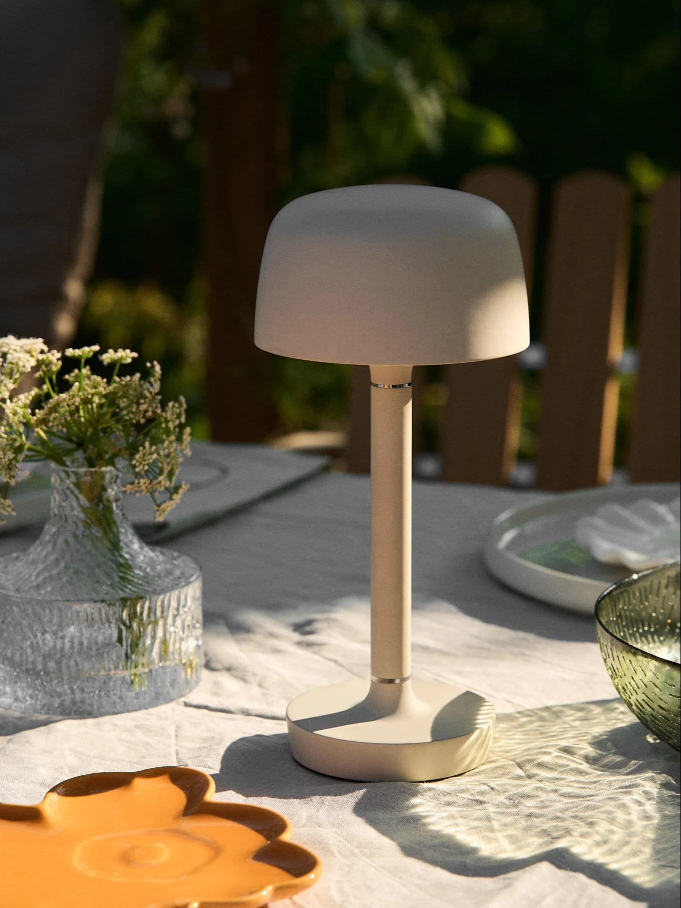 Beige portable table lamp on an outdoor table with a vase of flowers and dappled sunlight.