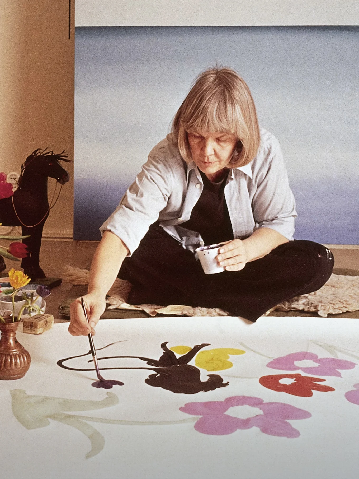 Artist Marimekko Maija Isola sitting on the floor painting large, colorful flowers on a white fabric.