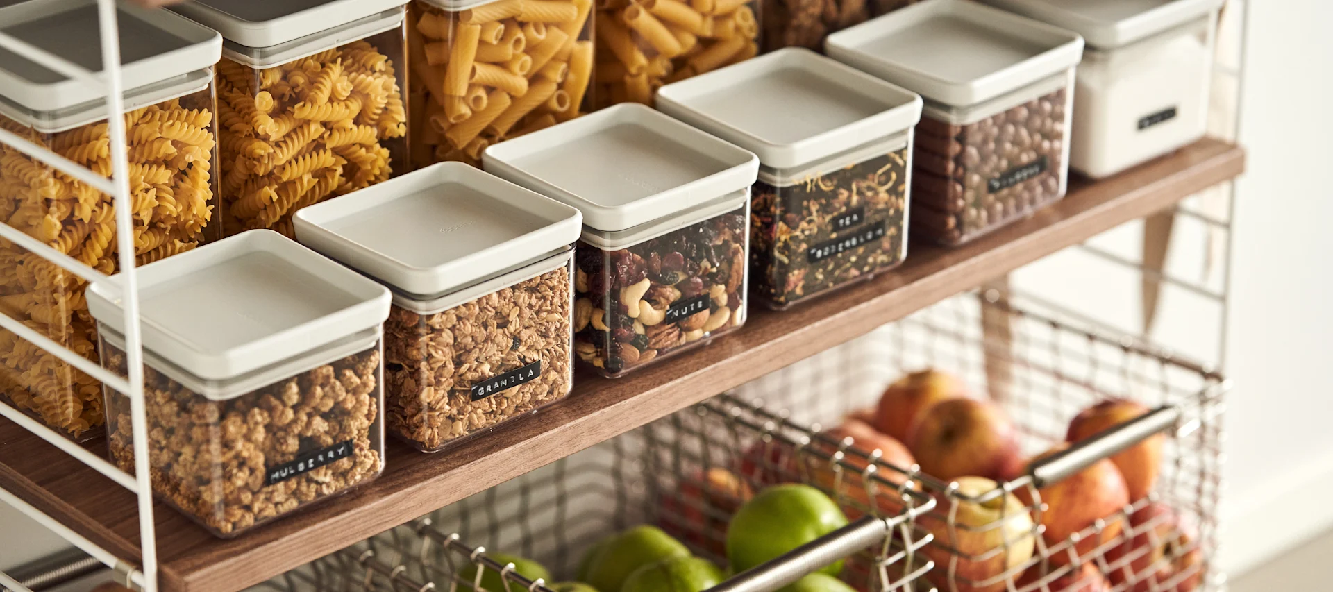 Close-up of a kitchen shelf with clear rectangular containers holding dry food: pasta, granola, nuts, and tea. Below, a wire basket holds apples.