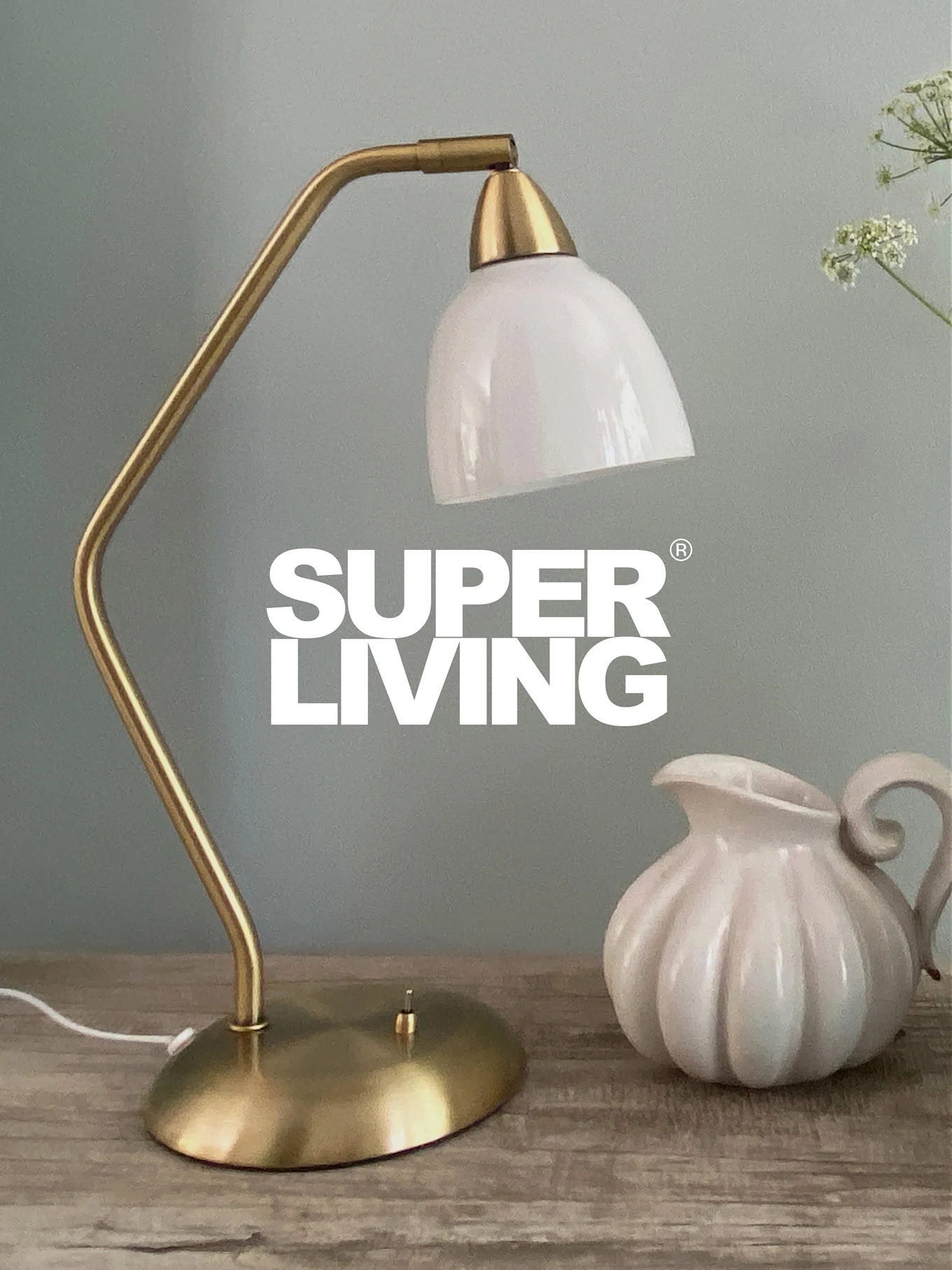 SUPER LIVING branding over a brass desk lamp with a white shade and a white, fluted pitcher on a wooden table.