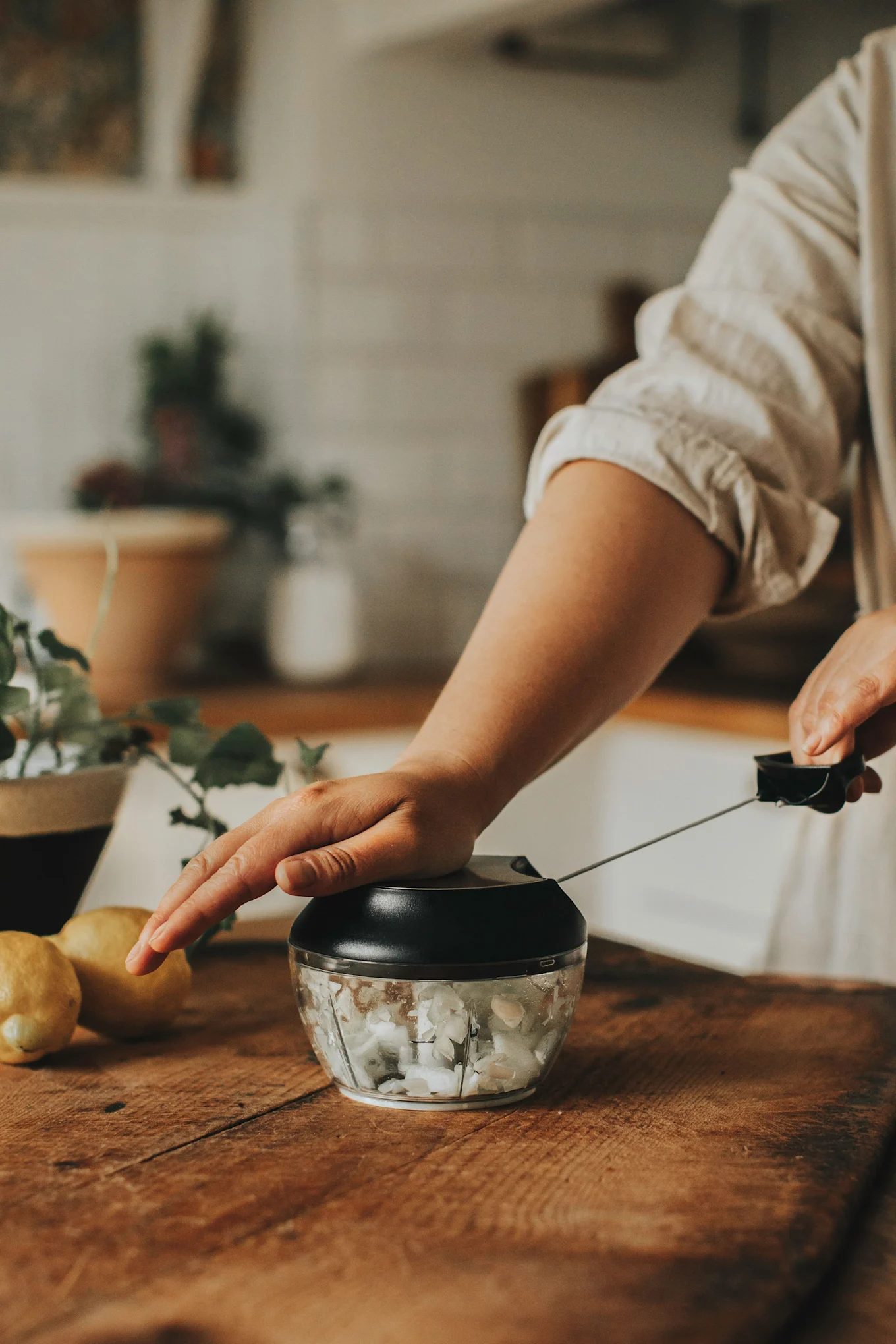 A person's hands operating a manual food chopper filled with white ingredients on a wooden counter with lemons.