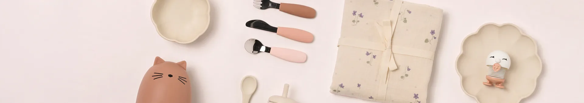 Baby dining essentials flat lay: beige plates, cat bottle, pink cutlery, floral blanket, egg toy.