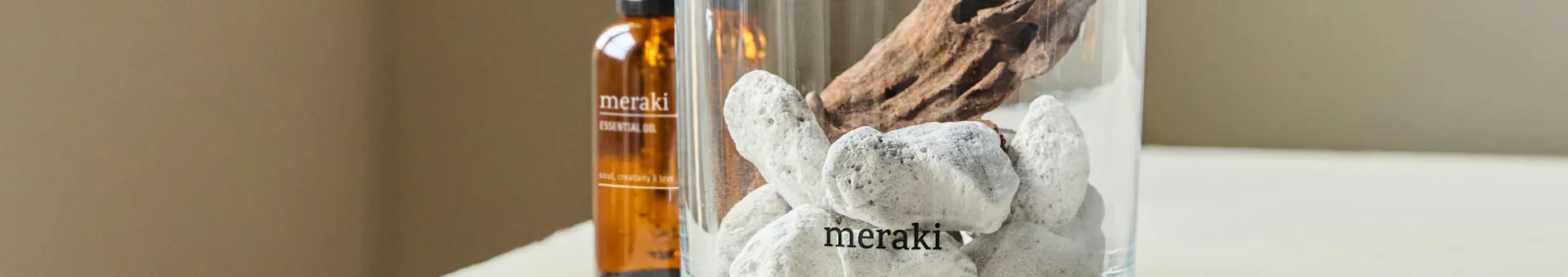 A brown essential oil bottle and a clear vase with white stones and driftwood. The bottle and vase are both labeled 'meraki'.