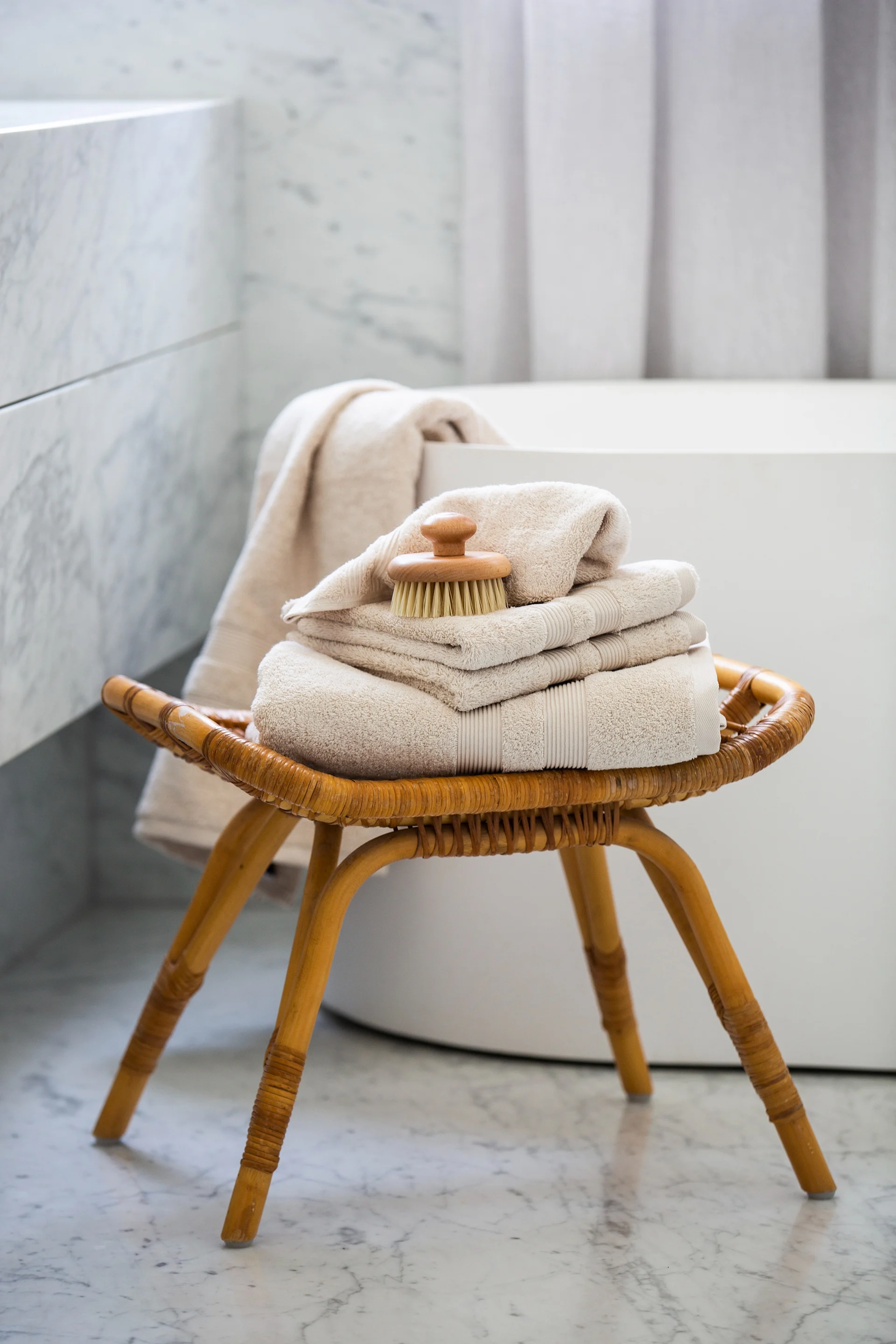 A rattan stool holds stacked beige towels and a wooden body brush in a marble bathroom.